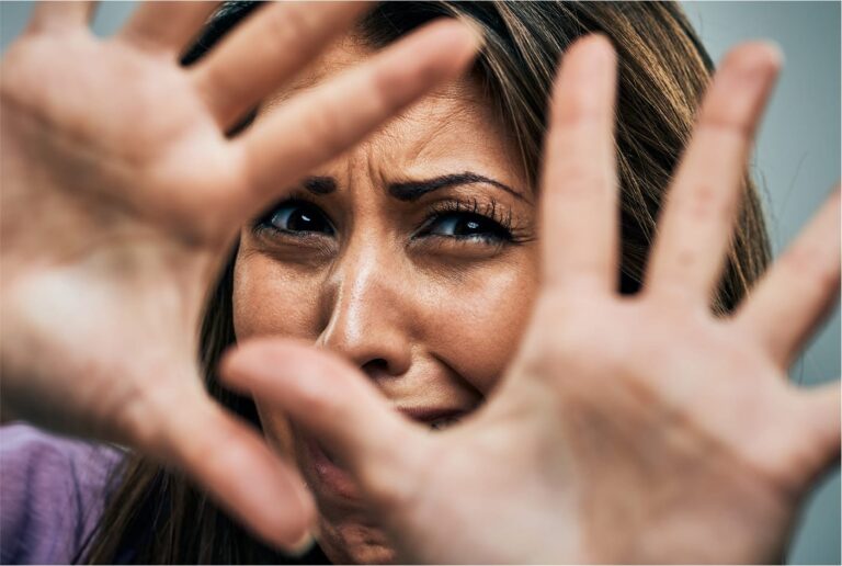 Frightened woman raising hands to shield herself, face centered between outstretched palms, symbolizing domestic violence and fear.