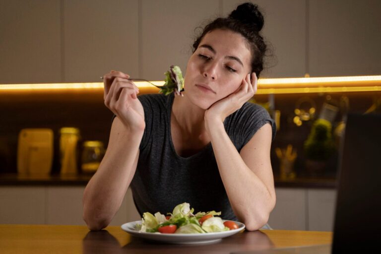 Woman sitting at a table holding a fork with salad, resting her head on her hand in a kitchen setting
