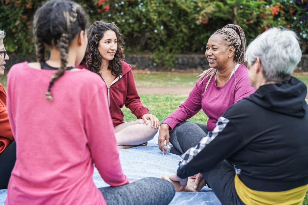 Group of women sitting in a circle on a blanket outdoors, engaged in conversation during a wellness or support group session in a park.