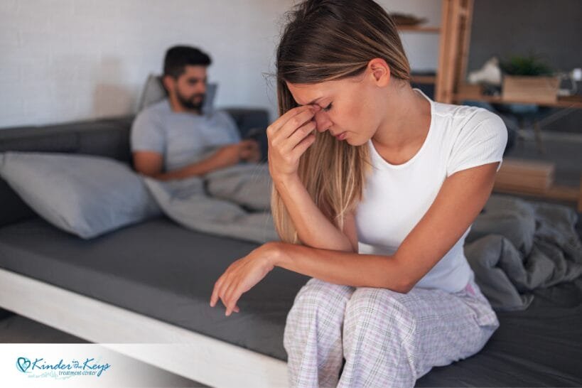 Woman sitting in bed feeling distressed while partner texts nearby